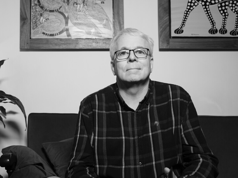 Older man wearing glasses and plaid shirt sitting on a dark couch indoors.