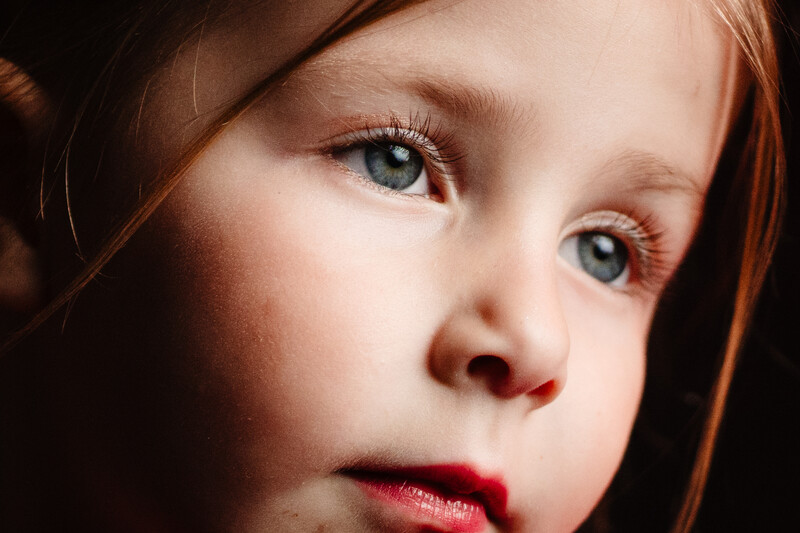 Close-up portrait of a young child with reddish-brown hair and blue eyes against a dark background.