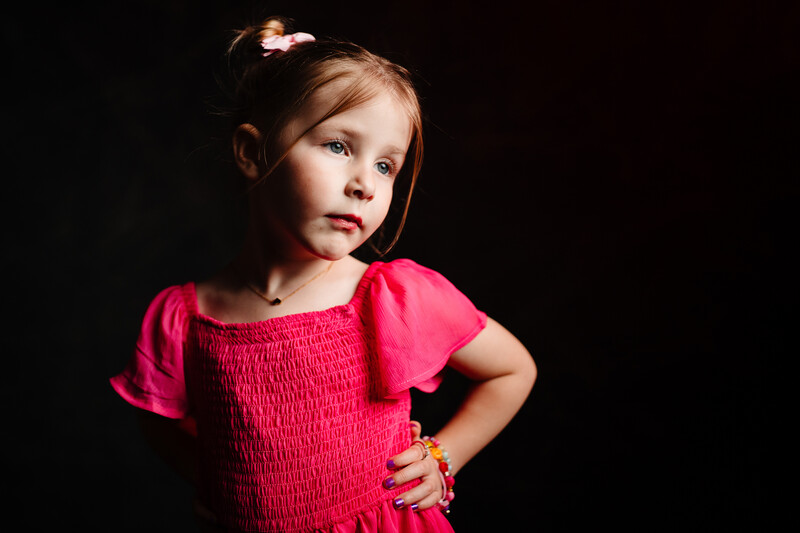 Young girl in bright pink dress with puffed sleeves against dark background.