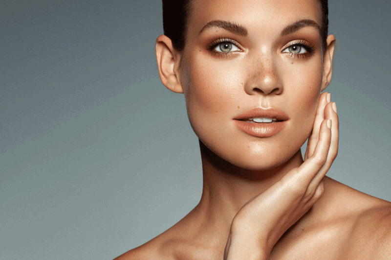 Woman with glowing skin looking at camera with hand touching her face against neutral background.