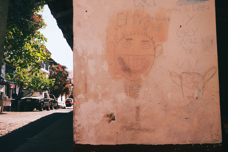 Terracotta-colored wall with child-like drawings and scratches, with sunlit street and ivy-covered building visible to the left.