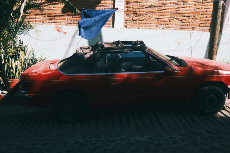 Red car with open roof and blue fabric suspended above it against a brick wall.