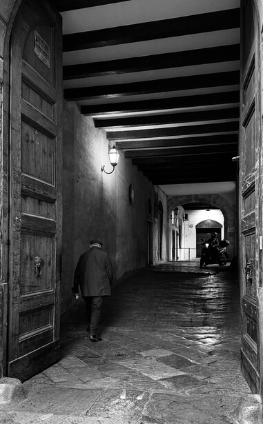 Person walking alone through a narrow, dimly lit alleyway with wet pavement and overhead beams.