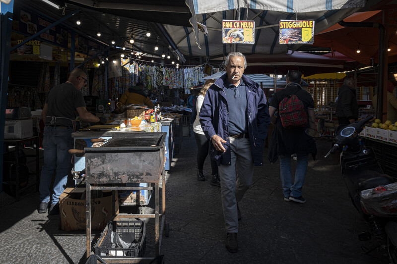 Man in blue shirt walking through covered market with food vendor stalls and hanging goods overhead.