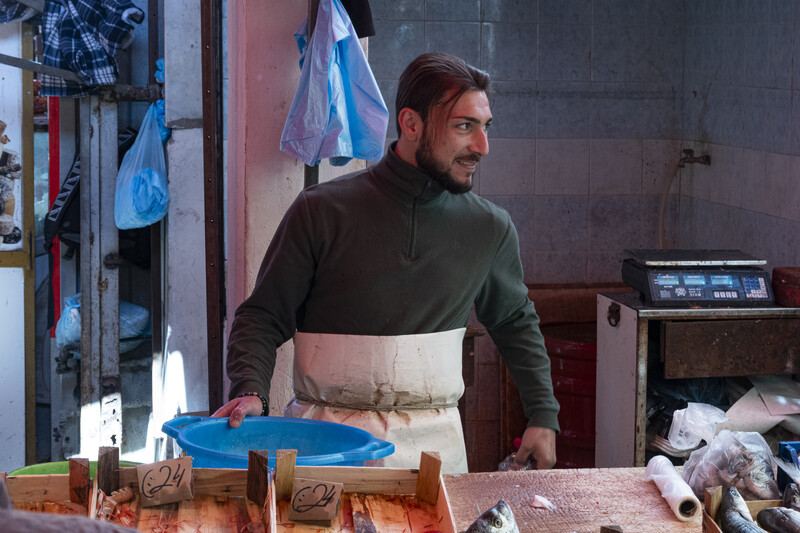 Man in olive and cream colorblock sweater and apron working at a wooden table in a workshop.