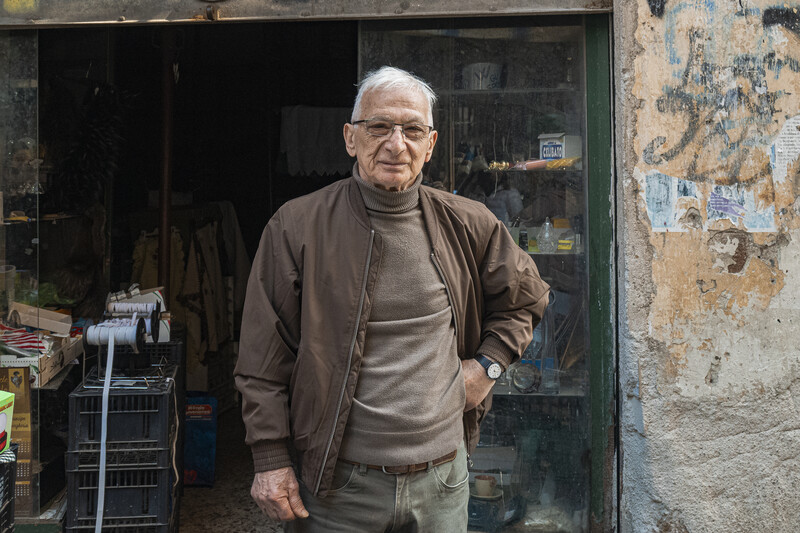 Elderly man with glasses standing in doorway of weathered storefront.