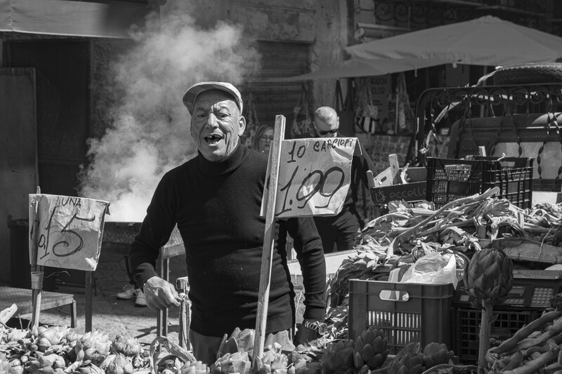 Man at a market stall with steam rising, holding a wooden pole near produce displays and handwritten signs.