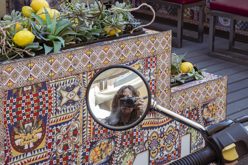 Magnifying glass reflecting a person's face held over ornately patterned decorative boxes with lemons and foliage.