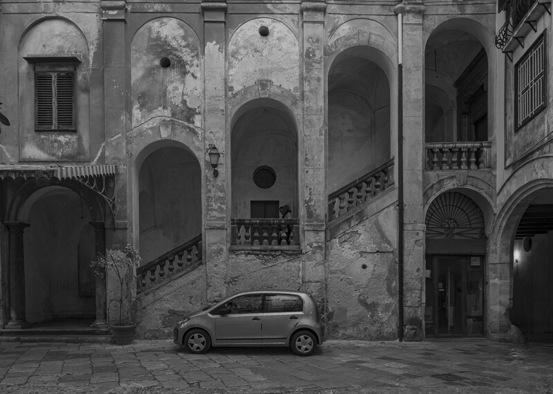 Gray compact car parked in a weathered courtyard surrounded by deteriorating classical architecture with arched doorways and ornate balconies.