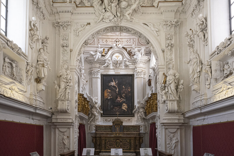 Ornate Baroque chapel interior with white sculptural reliefs, arched ceiling, and dark painting above altar.
