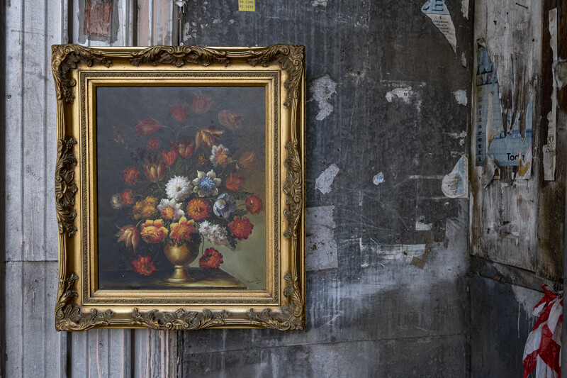 Ornate gold-framed still life painting of flowers in a vase hung on a weathered wall.