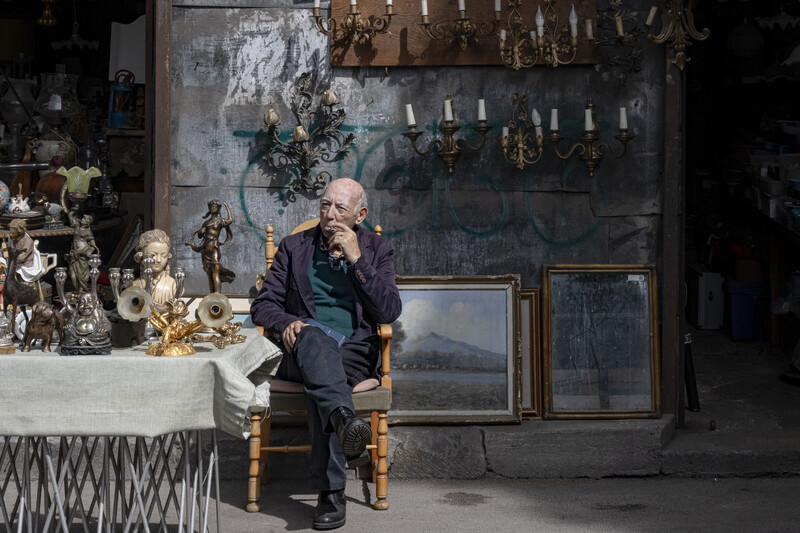 Elderly man in dark suit seated among antique sculptures and framed artworks in an ornate studio setting.