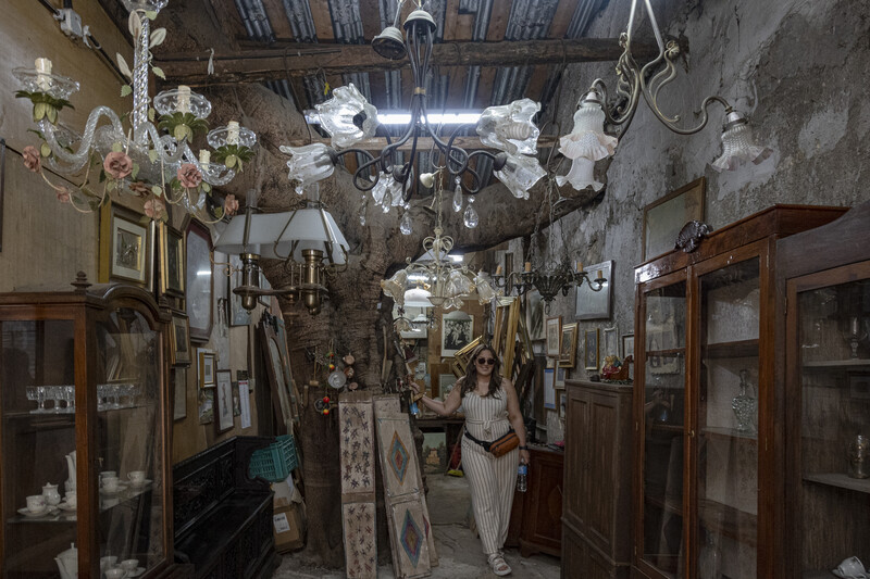 Woman standing in crowded antique shop corridor lined with wooden cabinets and vintage items.