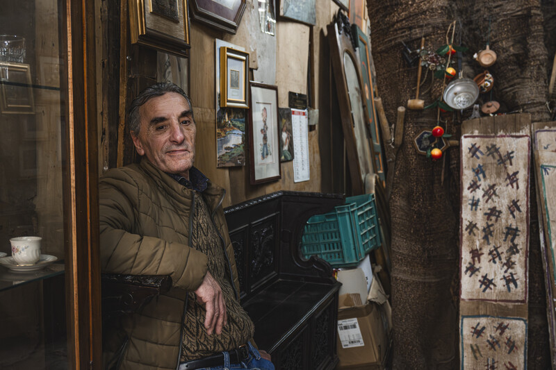 Man in olive jacket posing in a cluttered antique shop filled with framed artwork and vintage objects.