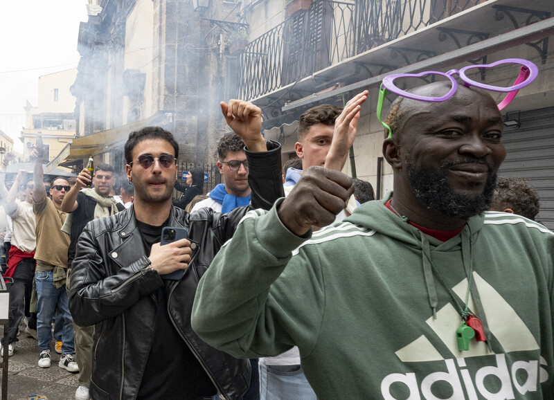 Street scene with people celebrating, one man in green Adidas hoodie raising his fist toward camera.