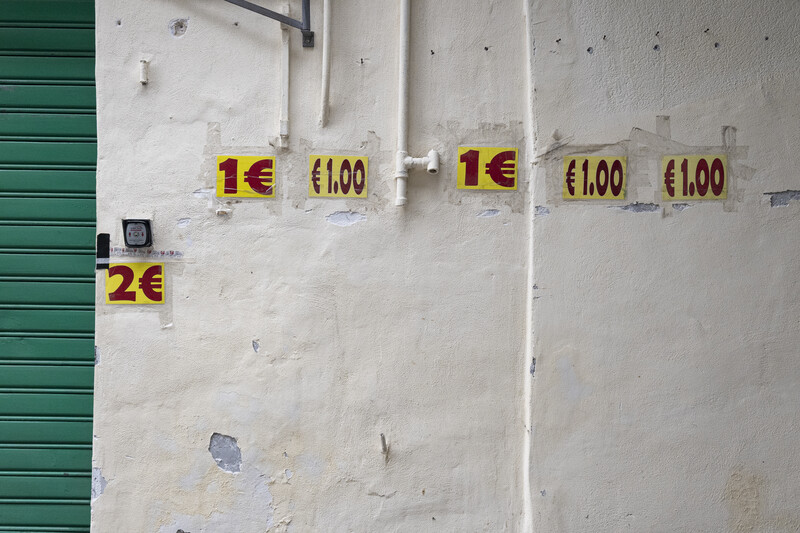 Price tags with euro amounts displayed on a weathered white wall next to a green metal shutter.