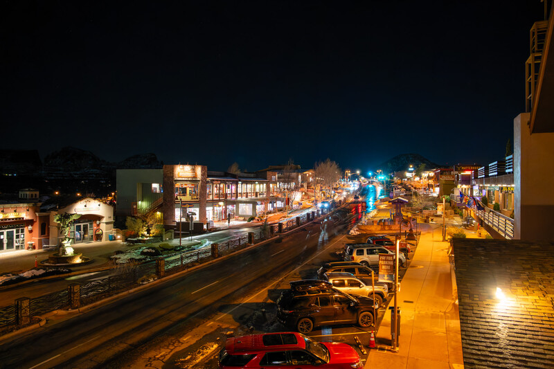 Elevated nighttime view of an urban street with illuminated storefronts, parked vehicles, and warm golden lighting under dark sky.