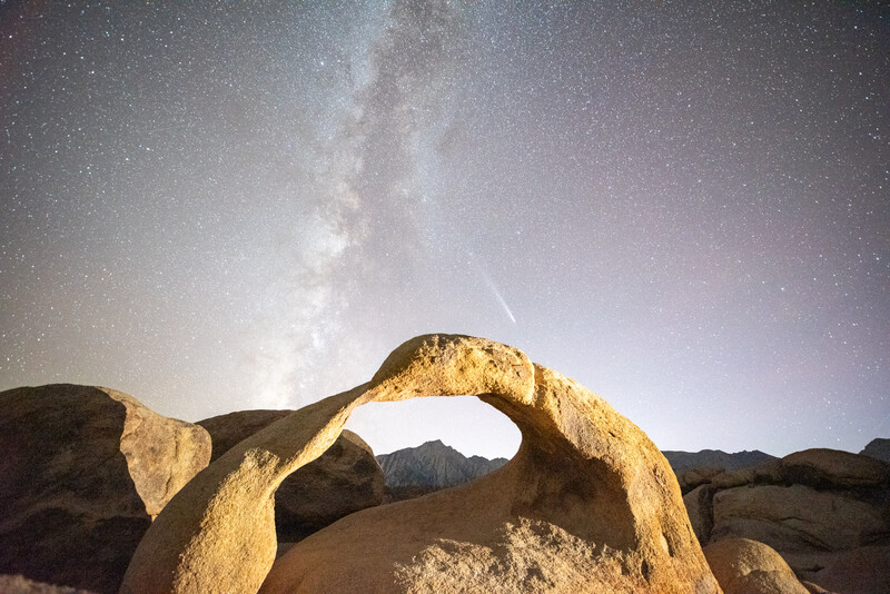 Natural rock arch framing the Milky Way galaxy and a meteor streak in a starry night sky.