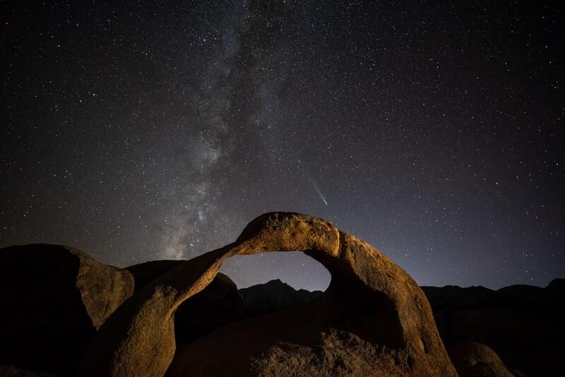 Natural rock arch framing the Milky Way galaxy and a meteor streaking across the night sky.