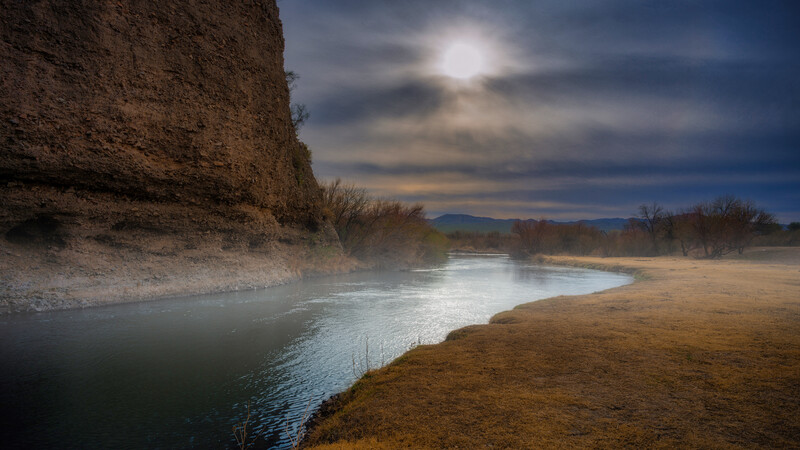 River winding through a valley beneath a tall rocky cliff under dramatic cloudy sky.