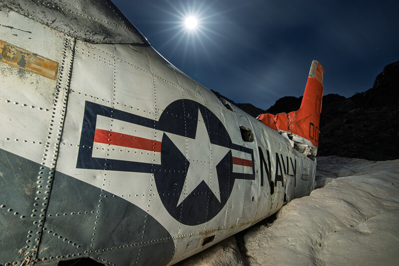Abandoned military aircraft fuselage with U.S. Navy star insignia on desert sand beneath dramatic sky.