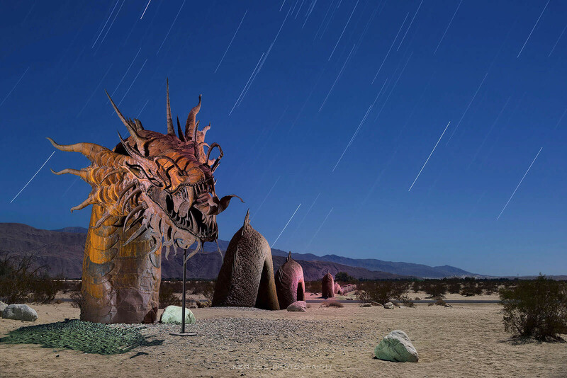 Large sculptural wooden head with extended spiky protrusions under star trails in desert landscape.