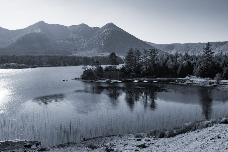 Snow-dusted mountains reflected in a partially frozen lake surrounded by evergreen forest.