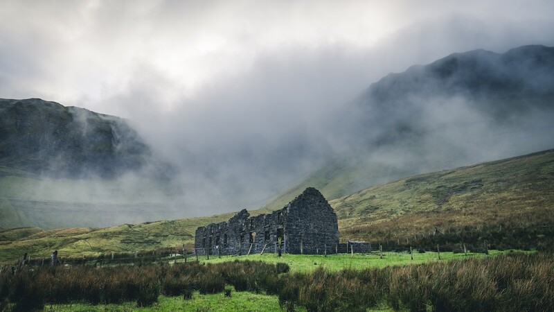 Stone ruins of a building standing in a misty valley beneath cloud-covered mountains.
