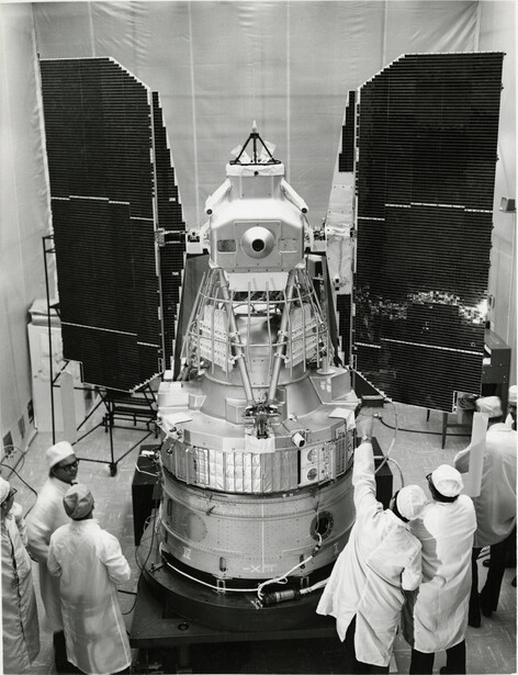 Spacecraft with deployed solar panels being examined by technicians in clean room environment.