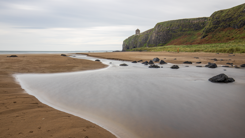 Tidal flat with shallow stream winding through sand toward moss-covered basalt cliffs under overcast sky.