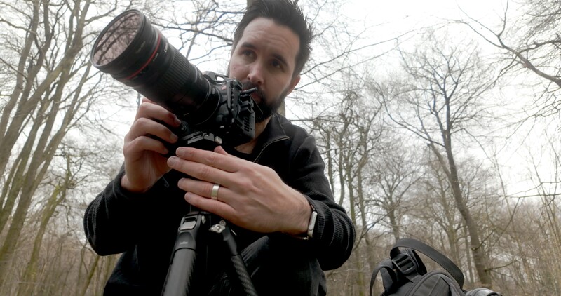 Photographer holding a DSLR camera with telephoto lens, looking down the barrel toward the camera in a wooded setting.