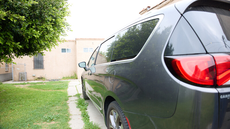 Dark minivan parked in residential driveway with overgrown grass and concrete pathway.