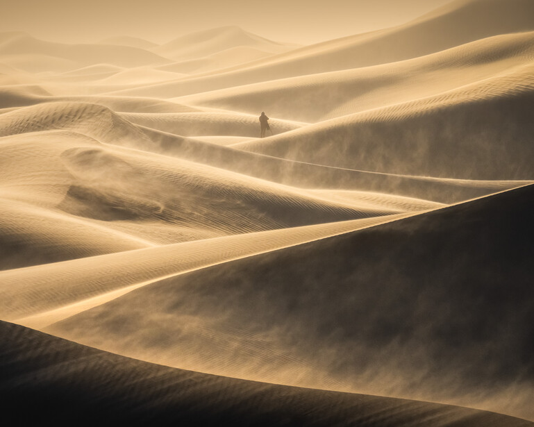 Solitary figure walking across windswept sand dunes in golden desert light.