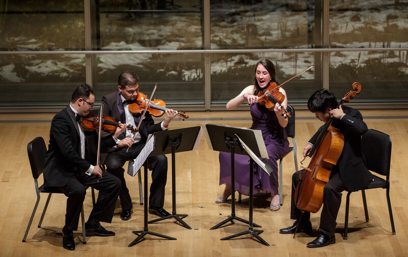 String quartet performing on stage with sheet music, seated in a semi-circle formation.