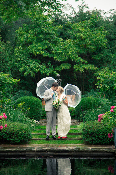Two people holding clear umbrellas stand by a pond with flowering shrubs and dense green foliage behind them.