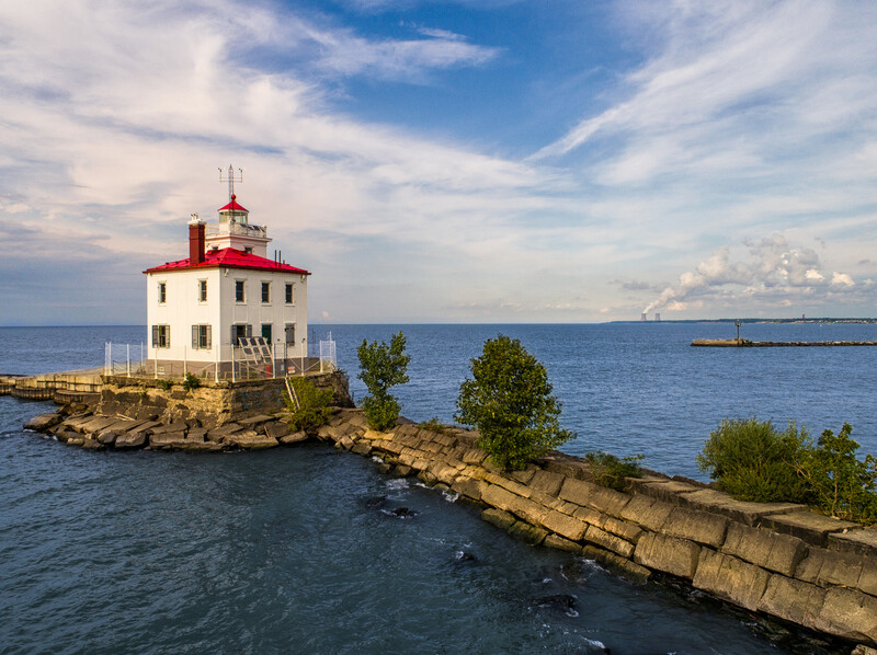 Red-roofed lighthouse on rocky outcrop surrounded by calm blue water under partly cloudy sky.