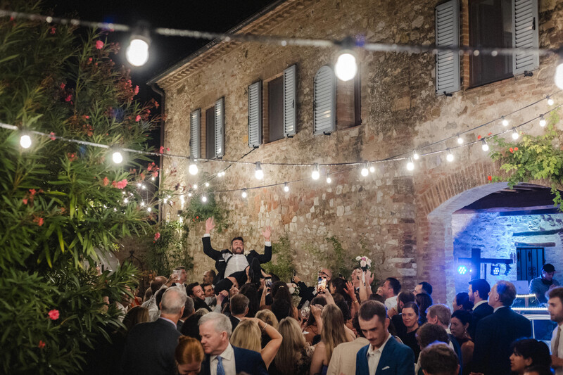 Crowd of formally dressed guests dancing and celebrating outside a rustic stone building at night.