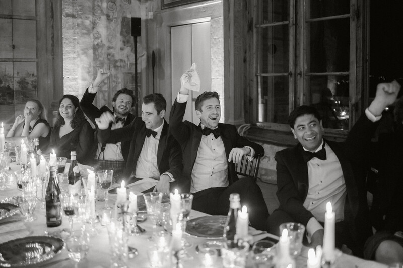 Men in formal wear seated at a long dinner table, celebrating with raised glasses and animated expressions.