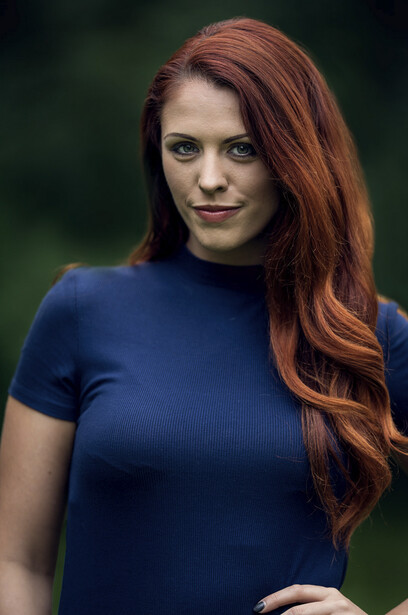 Woman with long reddish-brown hair wearing a dark blue short-sleeved shirt against a blurred green background.