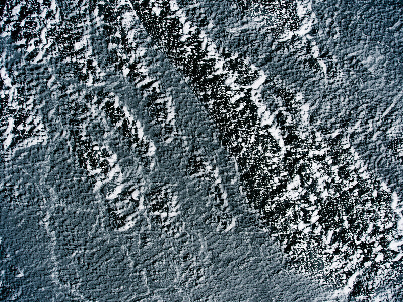 Aerial view of snow-covered mountain ridges with exposed dark rock faces creating diagonal striations across the frame.
