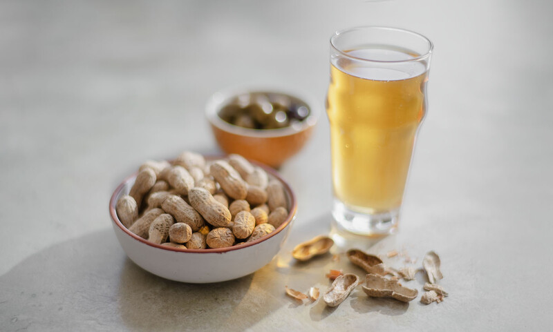 Glass of beer next to bowls of peanuts and olives on a gray surface.