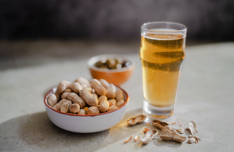 Glass of beer next to bowls of peanuts and olives on a concrete surface.