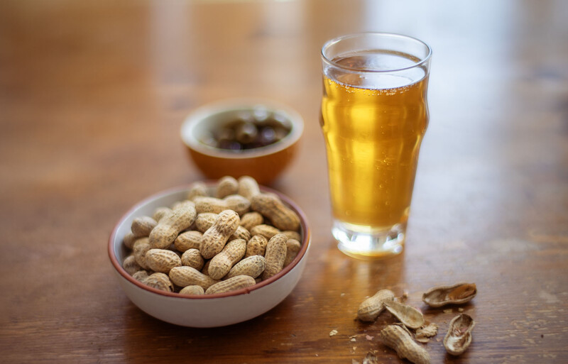Glass of beer next to a bowl of peanuts on a wooden surface.