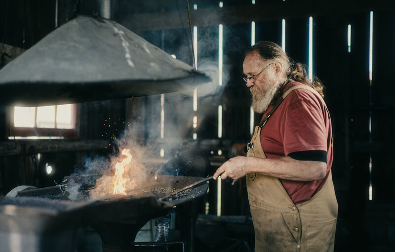 Blacksmith working at an anvil with hammer and glowing metal, illuminated by forge fire and overhead lights.