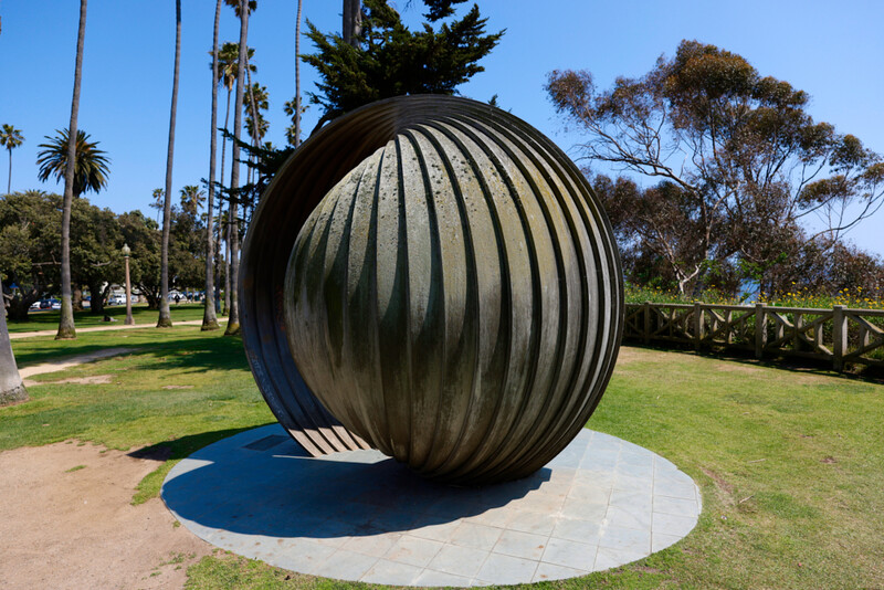 Large ribbed metal sphere sculpture installed on a circular concrete platform in a park setting.