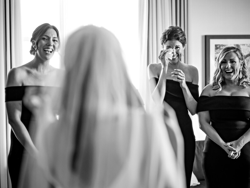 Bride in flowing veil smiles while bridesmaids in black dresses react emotionally behind her.