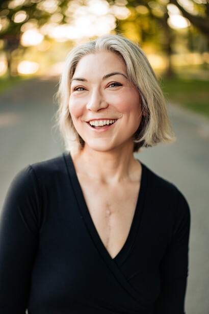 Portrait of a smiling woman with shoulder-length gray hair wearing a black v-neck top outdoors.