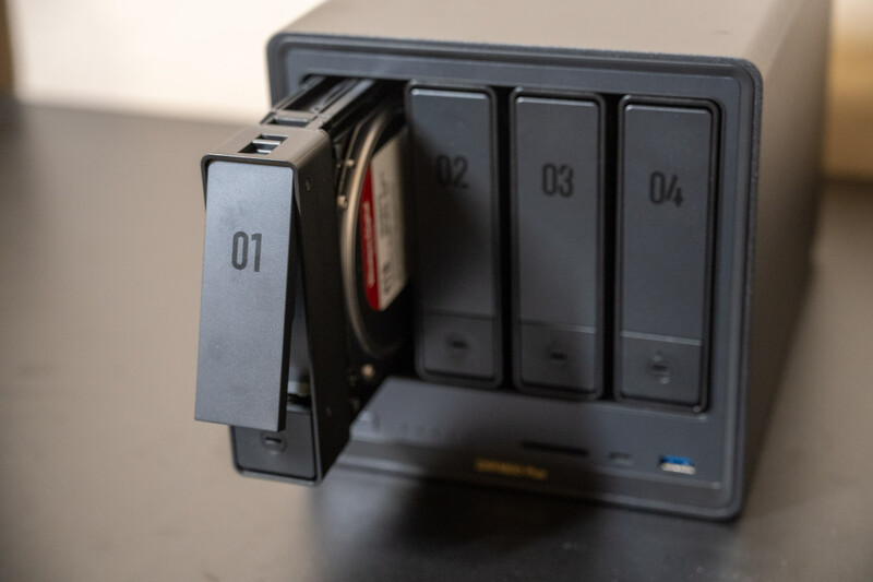 NAS storage enclosure with four drive bays, one hard drive partially inserted into the leftmost slot.