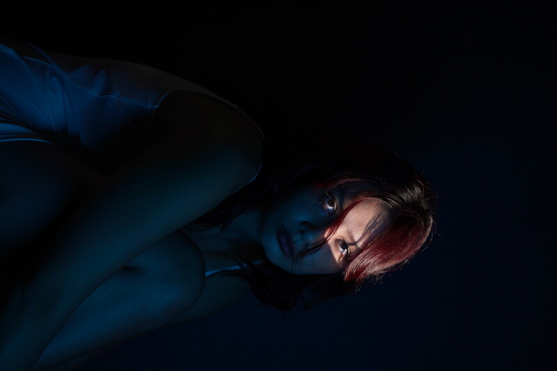 Person with dark hair and blue face paint against a black background, looking downward with dramatic side lighting.
