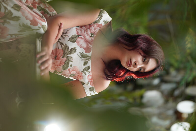 Woman with dark red wavy hair wearing a floral off-shoulder dress, gazing upward in an outdoor garden setting.